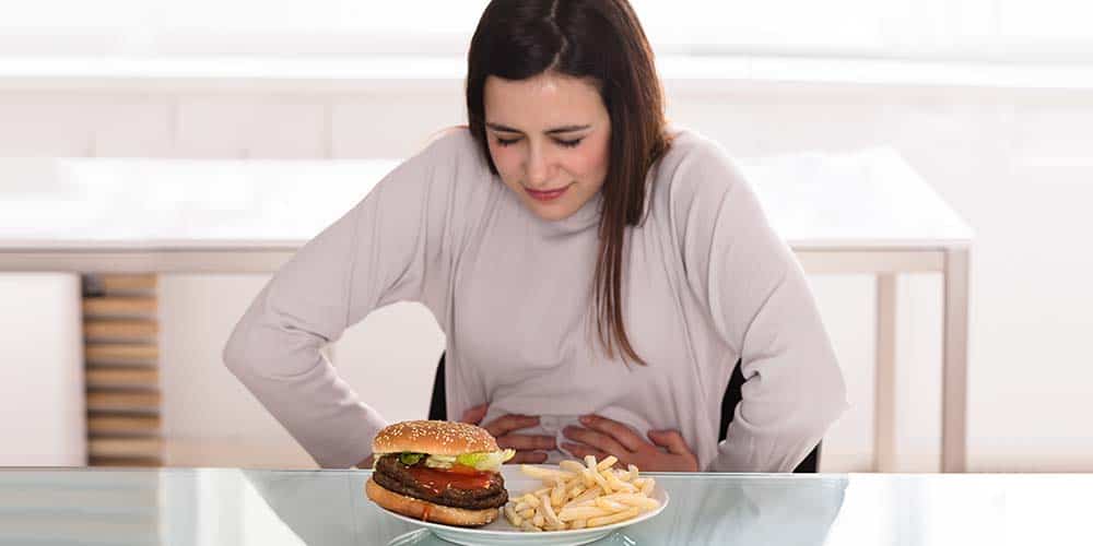 Junge Frau am Tisch hält sich den Bauch, vor ihr ein Teller mit Fast Food